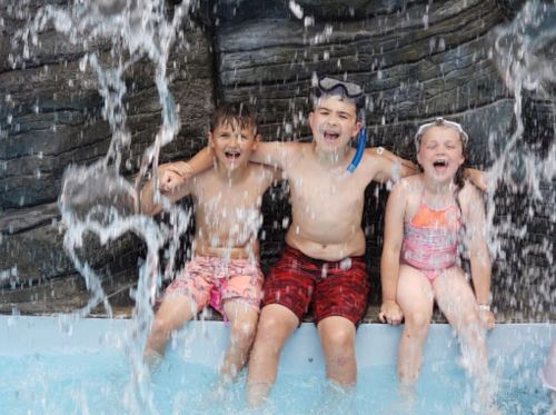 Three children sit together in a pool, laughing and enjoying a waterfall splashing over them.