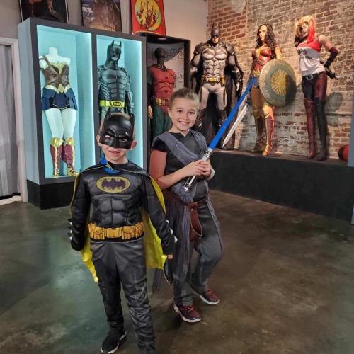 Two children dressed as Batman and Rey stand in a room with superhero displays in the background.