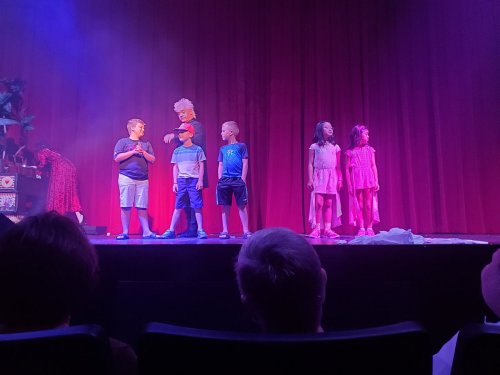 A group of children perform on stage with colorful lighting and a red curtain backdrop.