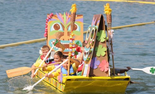 A colorful, decorated boat with children paddling, featuring tiki masks and floral decorations on a sunny waterway.