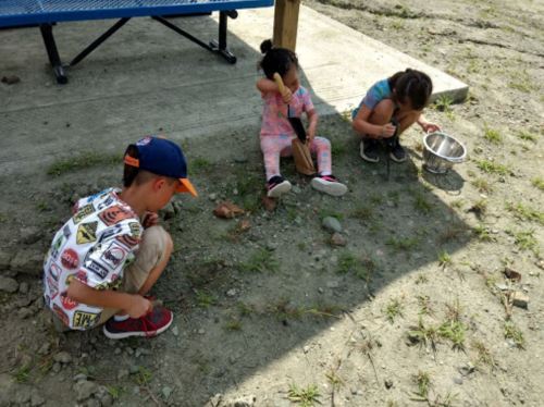 Three children are sitting on the ground, engaged in a creative activity with rocks and tools in a sunny outdoor setting.