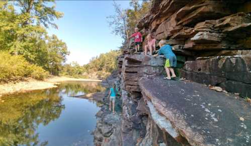 Children climbing on rocky terrain near a calm body of water, surrounded by trees and natural scenery.