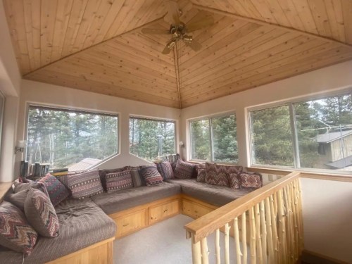 Cozy sunroom with a wooden ceiling, large windows, and a cushioned seating area surrounded by trees.