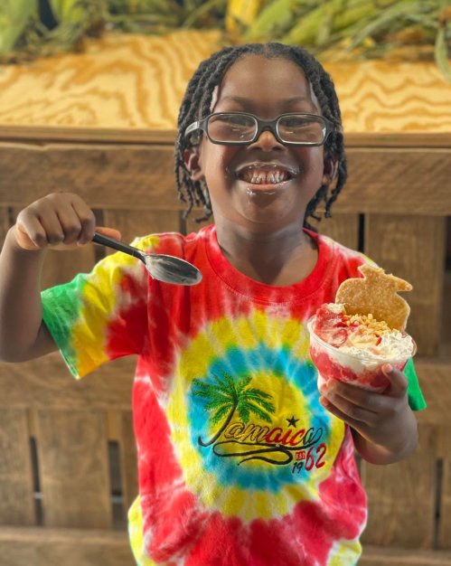 A smiling child in a colorful tie-dye shirt holds a dessert and a spoon, enjoying a sweet treat.