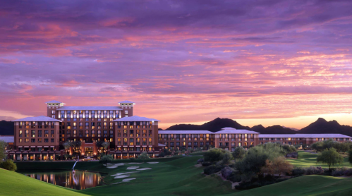 A luxurious hotel at sunset, surrounded by mountains and lush green golf courses under a colorful sky.