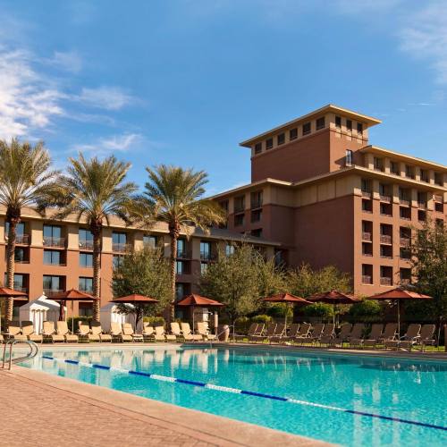 A sunny hotel pool area with lounge chairs, palm trees, and a large building in the background.
