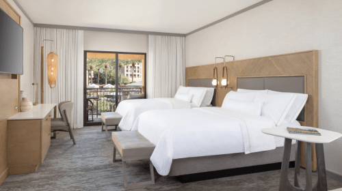 A modern hotel room featuring two beds, a desk, and a balcony with natural light and neutral decor.
