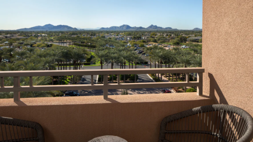 A balcony view overlooking palm trees and a valley with mountains in the distance under a clear blue sky.