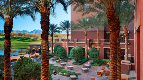 Lush courtyard with palm trees, seating areas, and a view of mountains in the background. Bright, sunny atmosphere.
