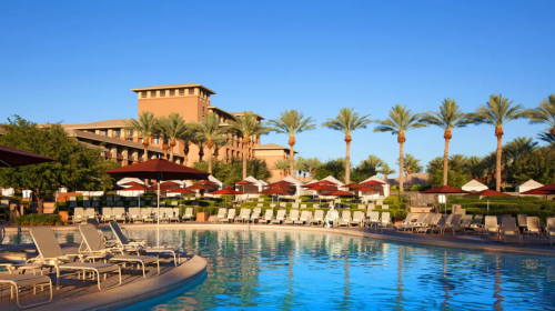 A sunny resort pool area with lounge chairs, palm trees, and a hotel in the background. Clear blue sky above.