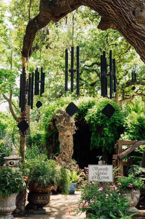 A lush garden with hanging black wind chimes, vibrant plants, and a welcoming sign amidst a large tree.