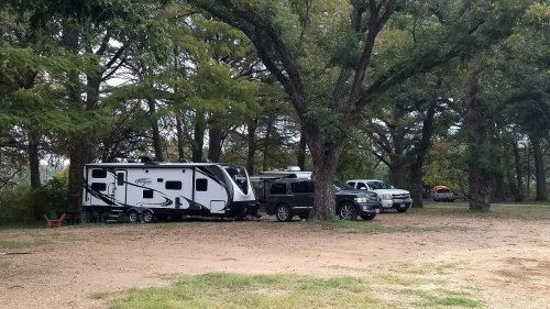 A white RV parked beside two trucks under large trees in a grassy area.