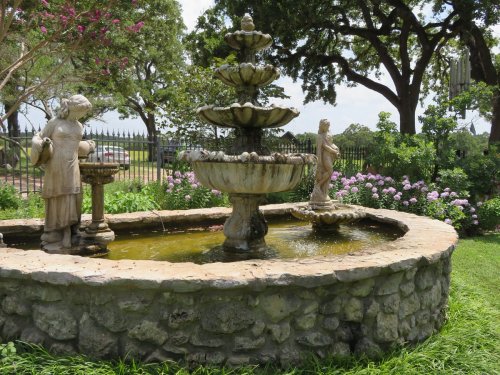 A stone fountain with two statues, surrounded by greenery and blooming flowers in a serene garden setting.