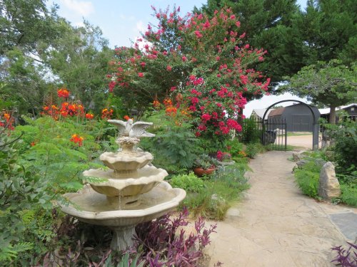 A stone fountain with a bird sculpture surrounded by vibrant flowers and greenery, leading to a garden gate.