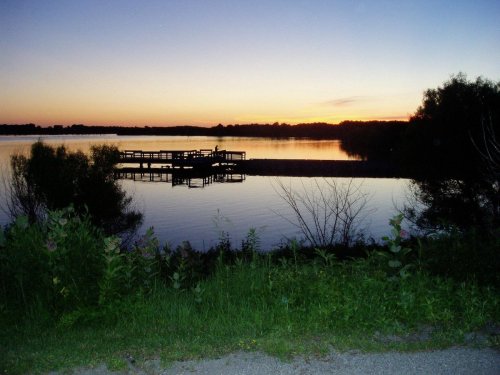 A serene lake at sunset, with a wooden dock and silhouettes of trees reflected in the calm water.