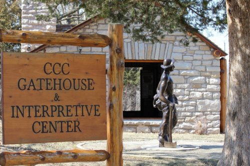 A stone building with a sign reading "CCC Gatehouse & Interpretive Center" and a statue of a man nearby.