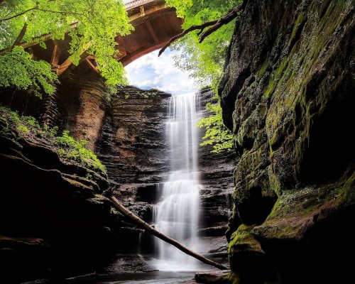 A serene waterfall cascades down rocky cliffs, framed by lush greenery and a bridge overhead.