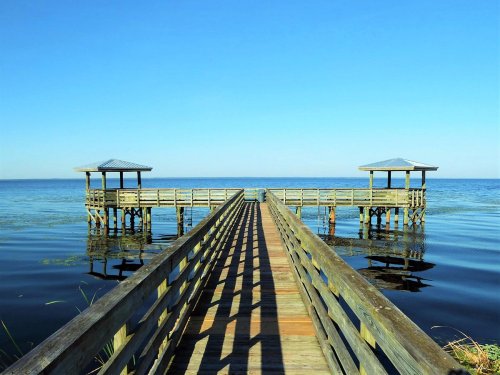 A wooden pier extends over calm water, leading to two gazebos under a clear blue sky.
