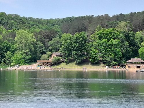 A serene lake scene with green trees, a sandy beach, and people enjoying the water on a sunny day.