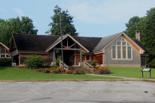 A modern wooden building with large windows, surrounded by greenery and flowers, set against a clear sky.