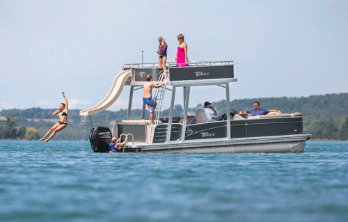 A group of people enjoying a pontoon boat on a lake, with one person jumping off a slide into the water.