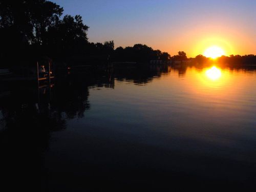 A serene sunset over a calm lake, with silhouettes of trees and docks reflecting on the water's surface.
