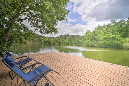 A serene lakeside view with a wooden dock and two lounge chairs, surrounded by lush greenery and cloudy skies.