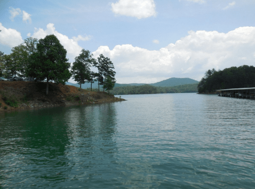 A serene lake surrounded by trees and mountains under a partly cloudy sky.