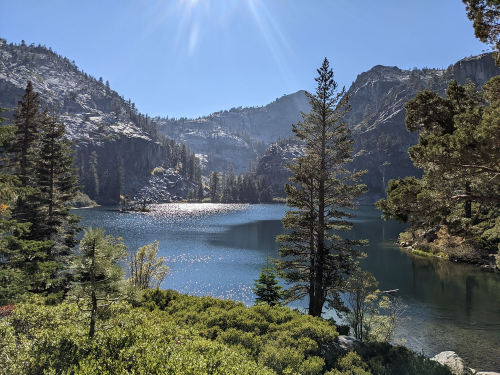 A serene lake surrounded by mountains and trees, reflecting sunlight under a clear blue sky.