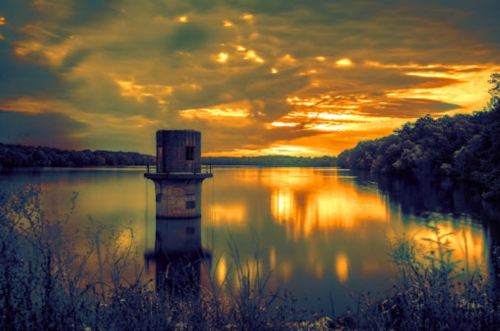 A serene lake at sunset, featuring a small tower on the water's edge, surrounded by trees and colorful clouds.