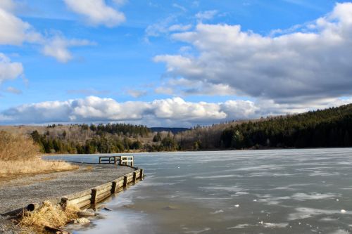 A serene winter scene of a frozen lake with a wooden pier, surrounded by trees and a cloudy blue sky.