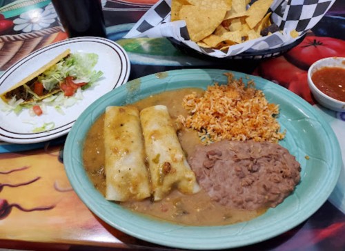 A plate of enchiladas with rice and refried beans, alongside a taco, tortilla chips, and salsa on a colorful table.