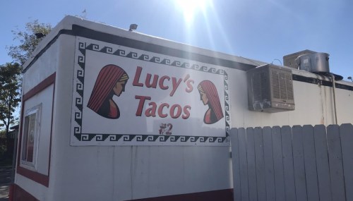 Sign for "Lucy's Tacos" on a food truck, featuring two stylized women and a bright sun in the background.