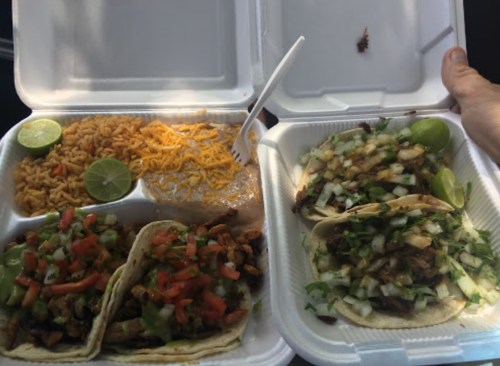 Two takeout containers with tacos topped with onions and cilantro, rice, and lime wedges.
