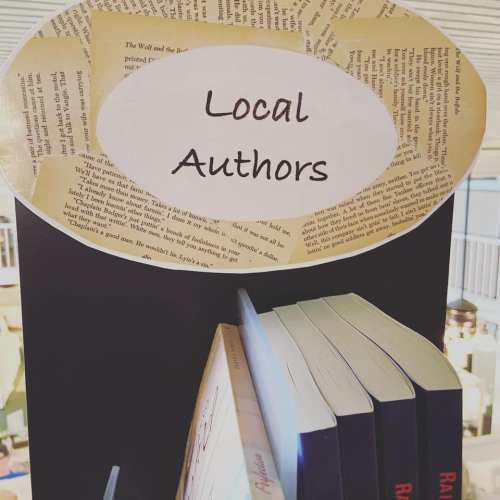 Sign reading "Local Authors" above a shelf of books, with pages from a book used as a decorative background.