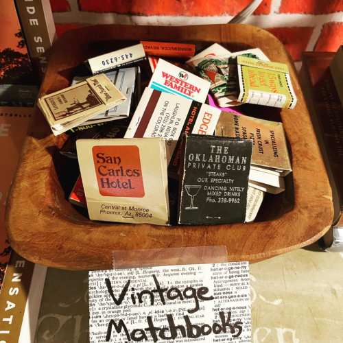 A wooden bowl filled with various vintage matchbooks, labeled "Vintage Matchbooks" on a sign.