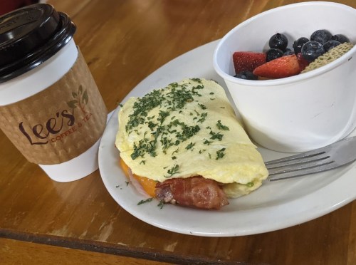 A plate with an omelet topped with herbs, bacon, and a side of fresh fruit, next to a coffee cup from Lee's Coffee.