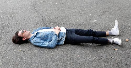 A person in a denim jacket and sunglasses lies on a paved surface, arms crossed, looking relaxed.