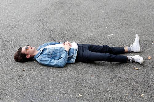 A person in a denim jacket and sunglasses lies on the pavement, hands on their stomach, looking relaxed.