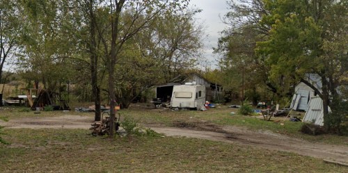 A rundown area with an old RV, dilapidated buildings, and scattered debris among trees and overgrown grass.