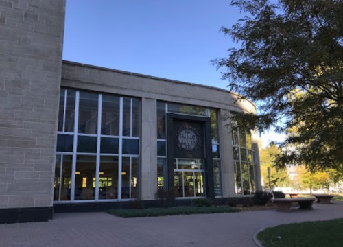 A modern building with large windows and a circular sign, surrounded by trees and a clear blue sky.