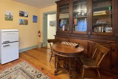 A cozy room with a white fridge, wooden table and chairs, a bookshelf, and framed pictures on the walls.