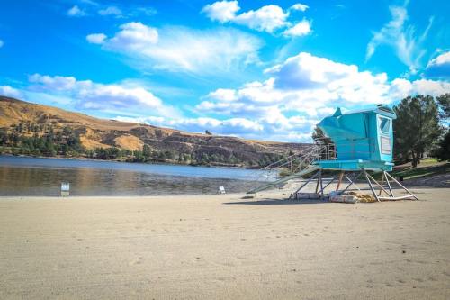 A blue lifeguard tower on a sandy beach by a calm lake, surrounded by hills and a clear blue sky with fluffy clouds.