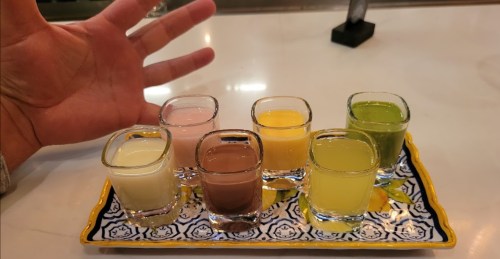 A hand gestures next to a tray of seven colorful shot glasses filled with various drinks.