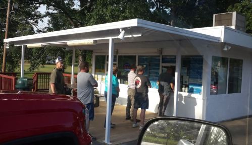 A group of people standing in line outside a small building with large windows, surrounded by trees.