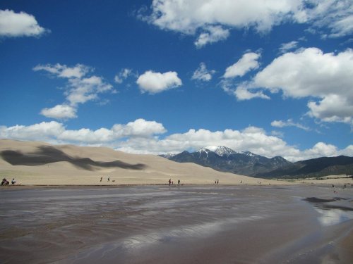A vast desert landscape with sand dunes, mountains in the background, and a blue sky dotted with fluffy clouds.