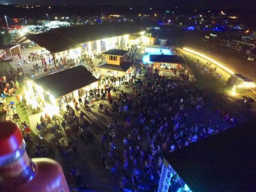 Aerial view of a large crowd gathered at a nighttime outdoor event, with illuminated buildings and a pool in the background.