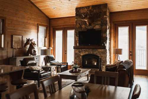 Cozy living room with stone fireplace, wooden walls, leather chairs, and large windows overlooking nature.