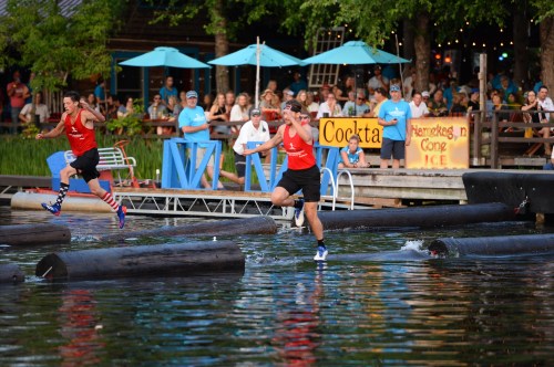 Two competitors race across floating logs on a lake, with spectators watching from the shore. Colorful umbrellas in the background.