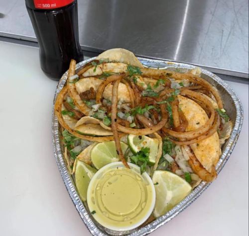 A heart-shaped tray of tacos topped with onions and cilantro, served with lime wedges and a small container of sauce.